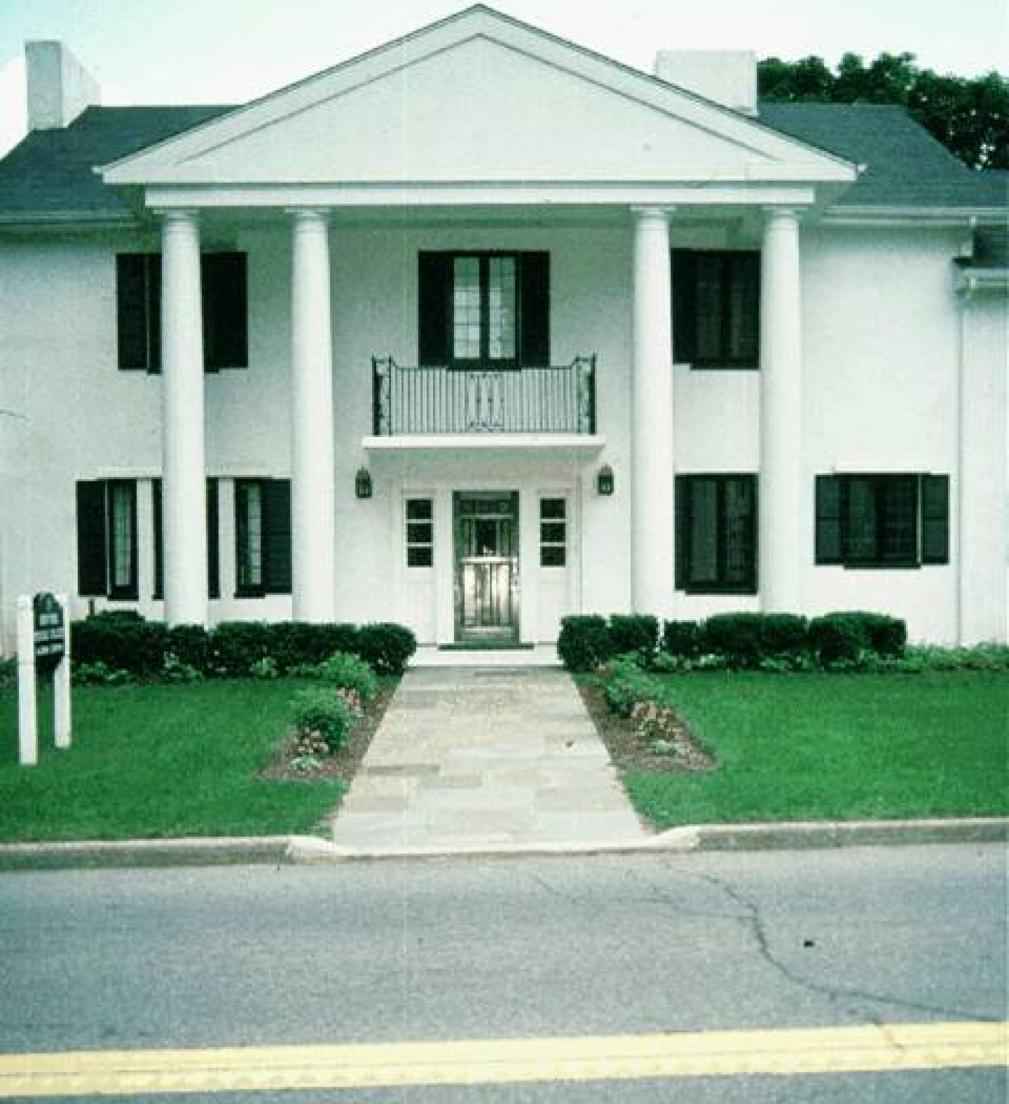 The alumni home, a two-story white house with large pillars, black shutters, a stone path, green bushes, and a green front yard