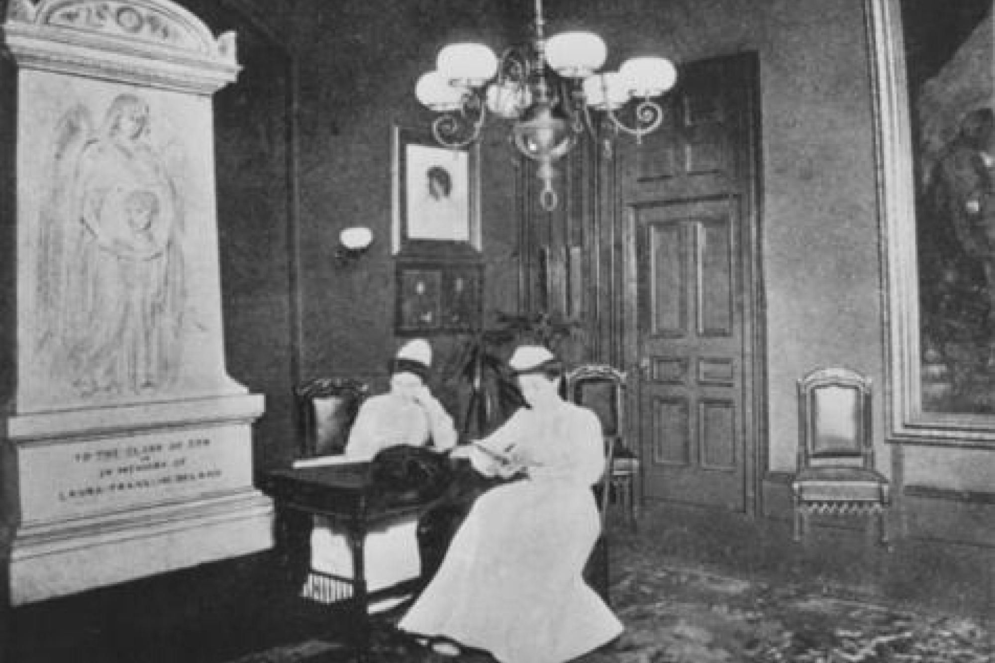 Two female nurses sitting in large room with large art, a chandelier, and a tall wood door at Fifth Ave Hospital.