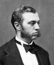 Ira Remson. A young looking man with dark, combed back hair, and mutton chops. He is wearing a suit and bowtie and is looking away from the camera