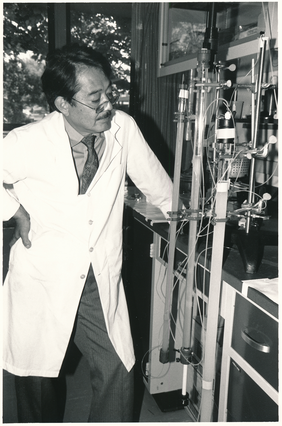 Kikkawa Yutaka, with dark hair, small glasses, and a lab coat with a button down shirt and tie under it. He is looking at his research equipment in the lab.