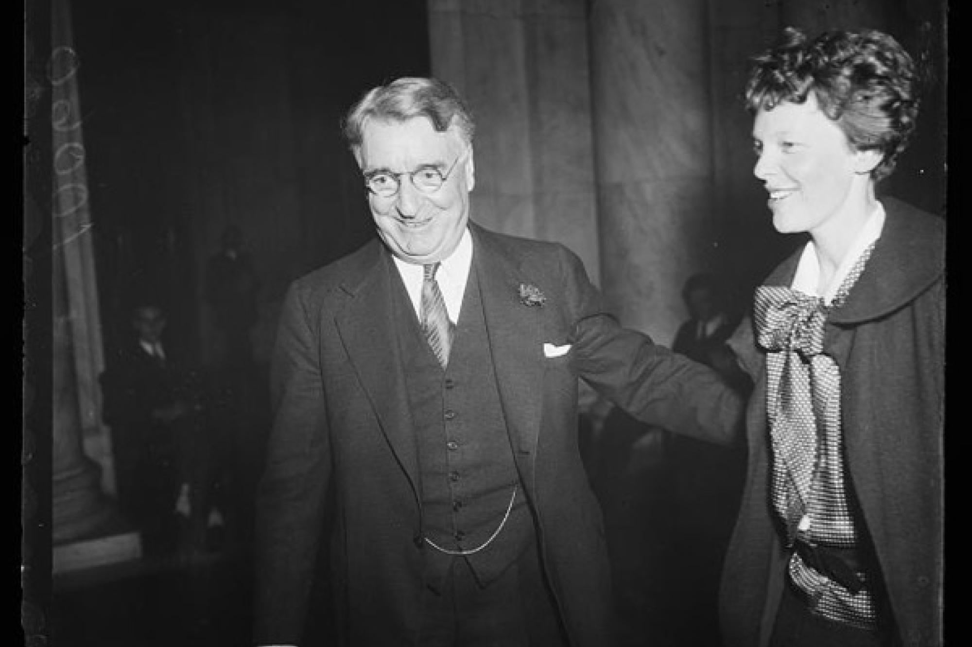 Royal S. Copeland, wearing a suit and circular glasses, smiling next to Amelia Earhart, wearing a patterned blouse and a jacket with a round collar