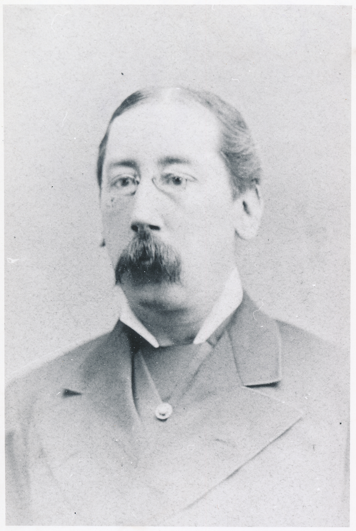 Portrait of Dr. Adolfo Varona, with short hair, small metal glasses, a thick mustache that covers his mouth, a white collared shirt that covers his neck, and a blazer.