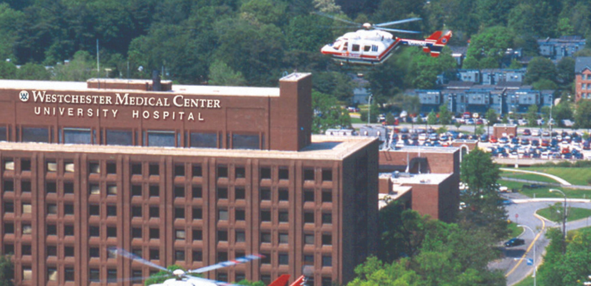 WMC, a large brick building with green trees, grass, and large parking lot in background. Helicopter is landing at hospital