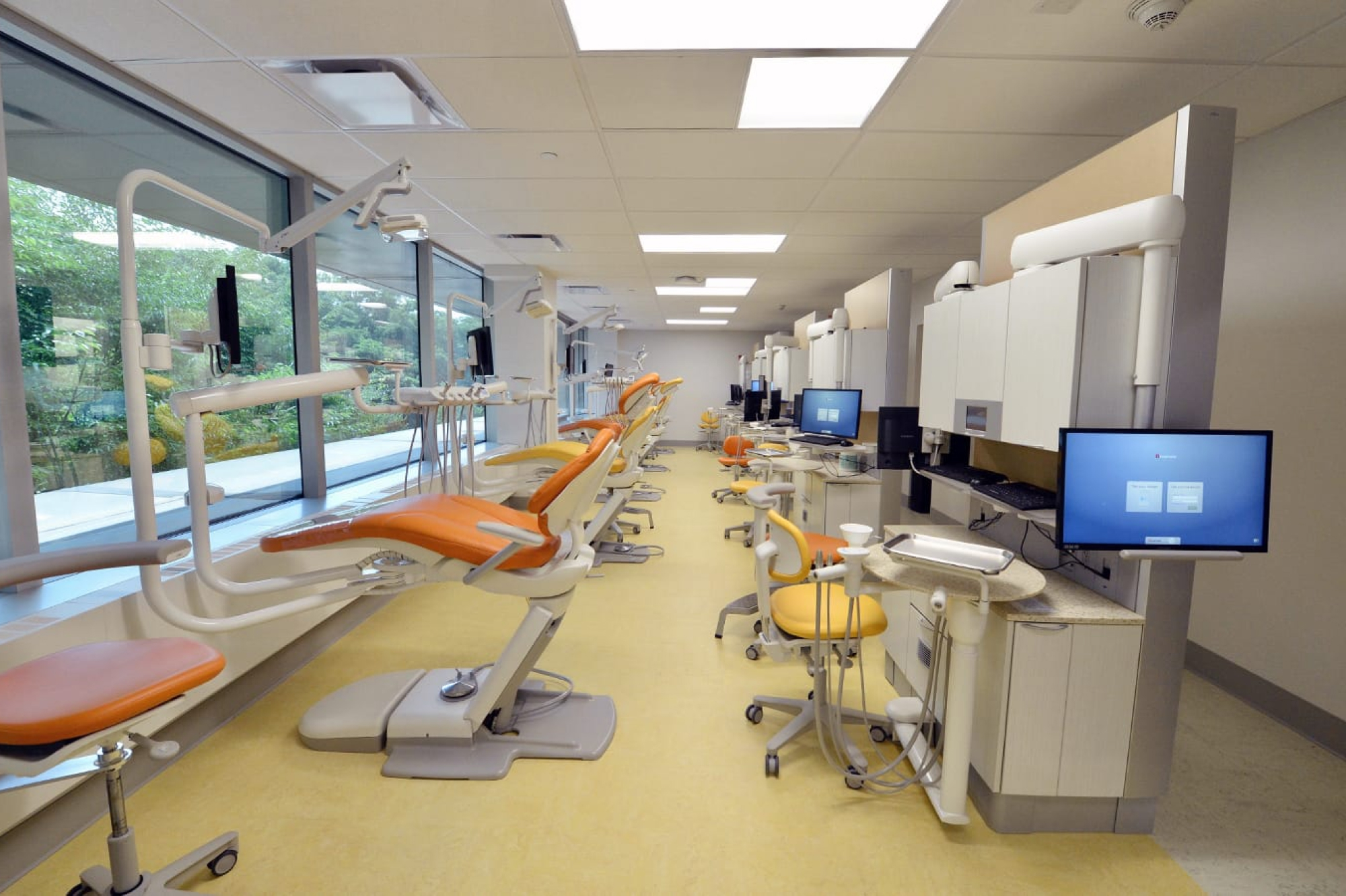 Line of orange dental chairs along window in clinic with computers across from them