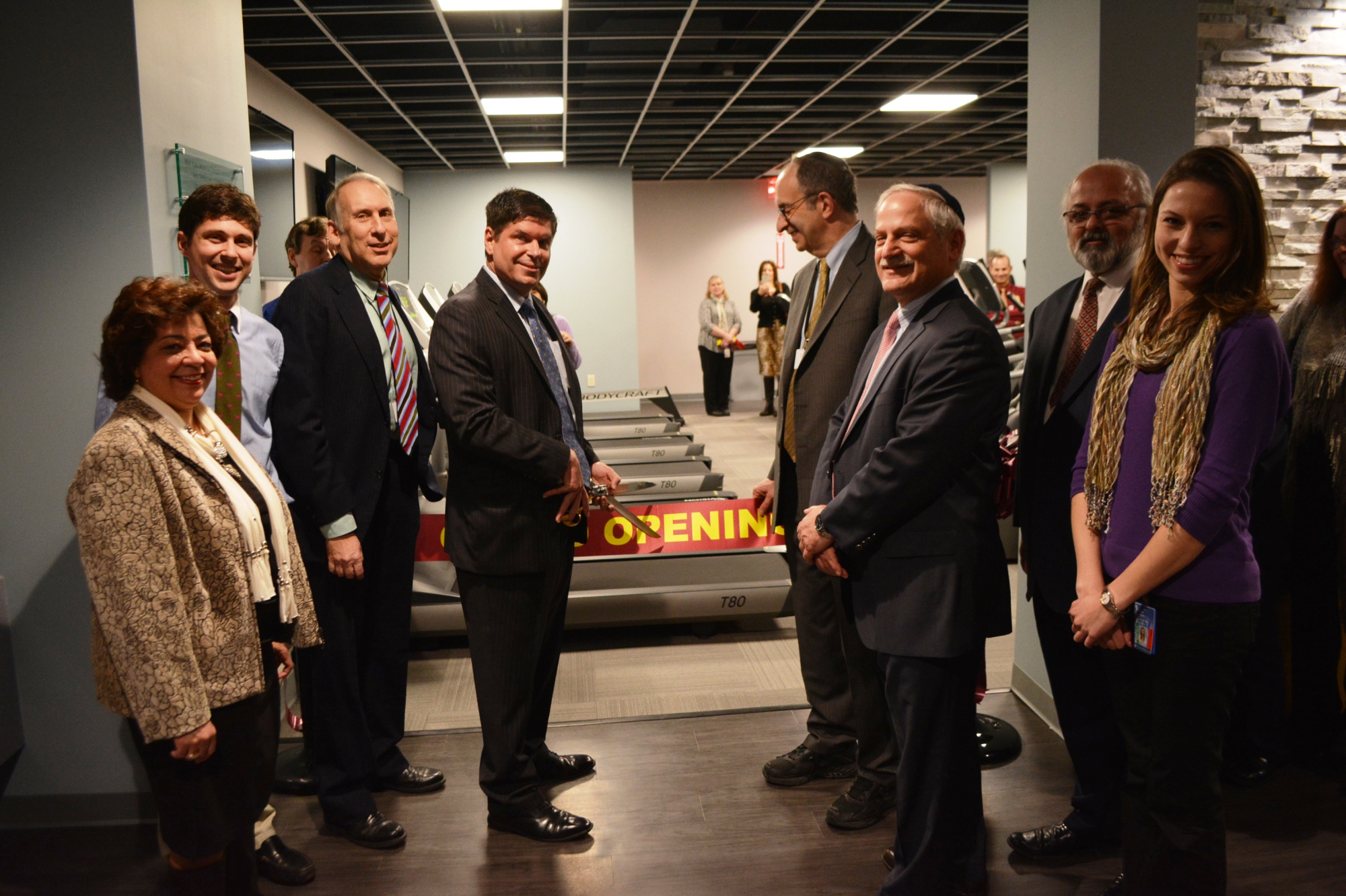 NYMC faculty and staff smiling next to grand opening ribbon at the fitness center with row of treadmills in background