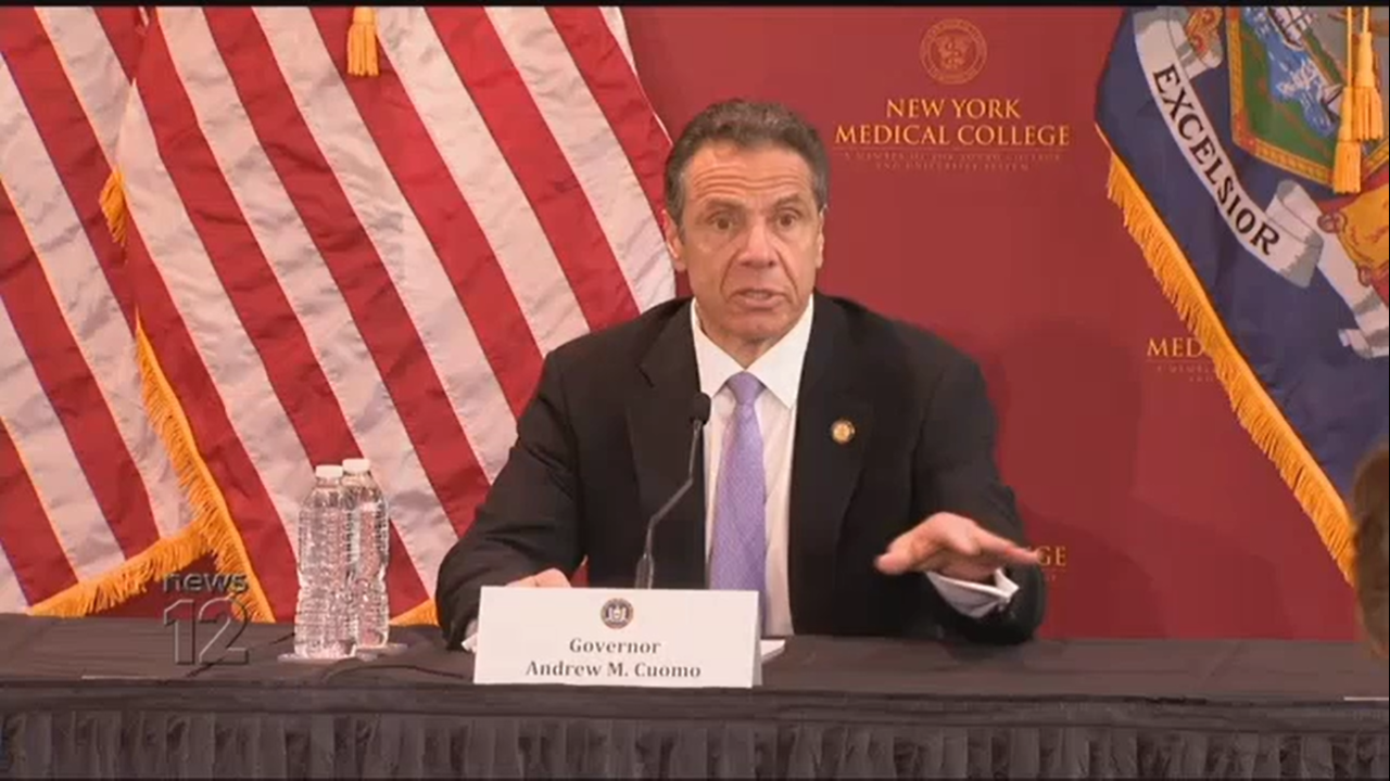 Governor Cuomo doing press briefing at NYMC with NYMC logo in background, an American flag, and a NYS flag