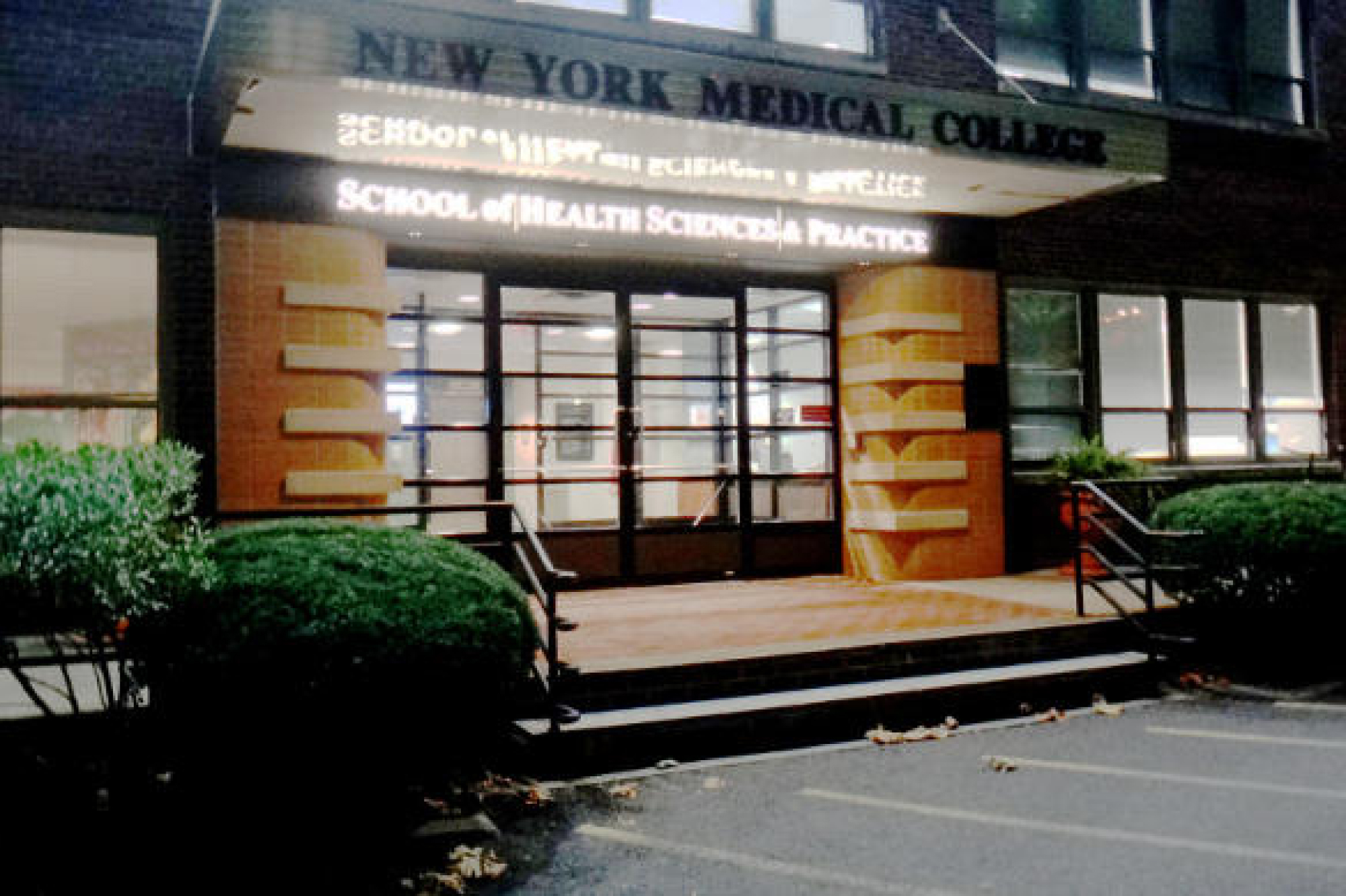 School of health sciences and practice exterior with an illuminated sign, many windows, and steps and bushes outside the building