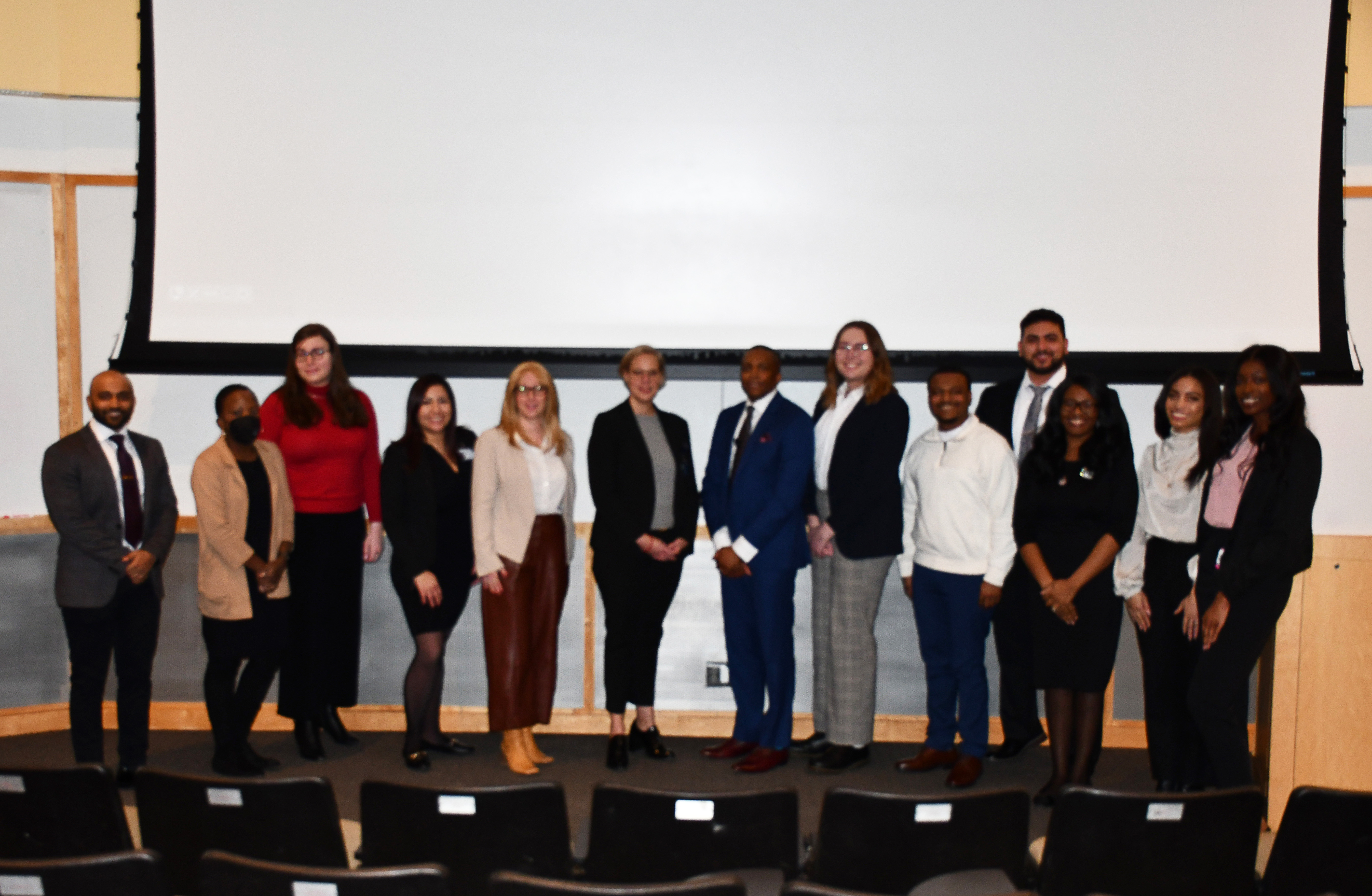 Leadership, students, faculty, and community members dressed in professional attire smiling in an auditorium