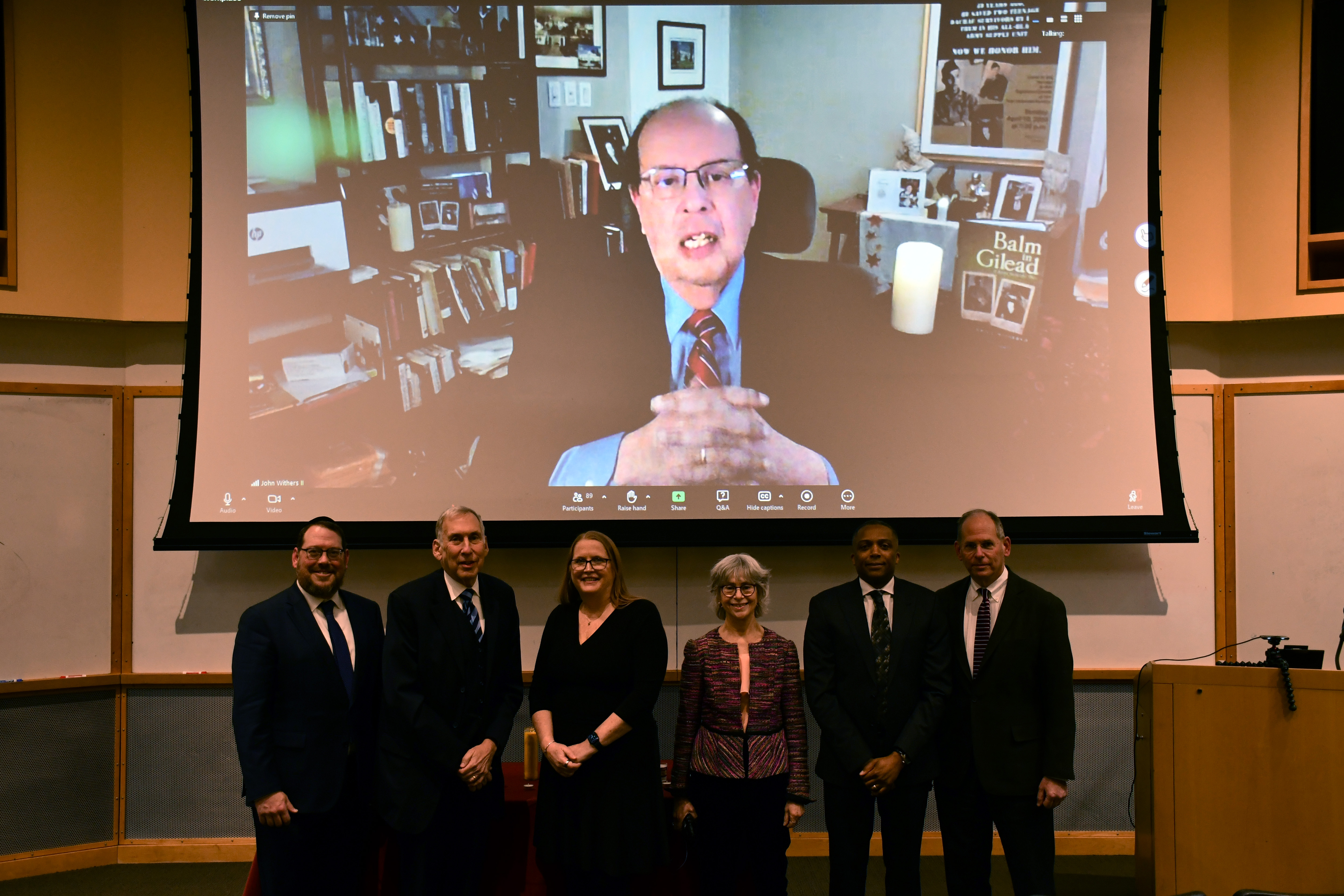 Men and women in an auditorium smiling, with a projector overhead displaying an image of a man in an office.