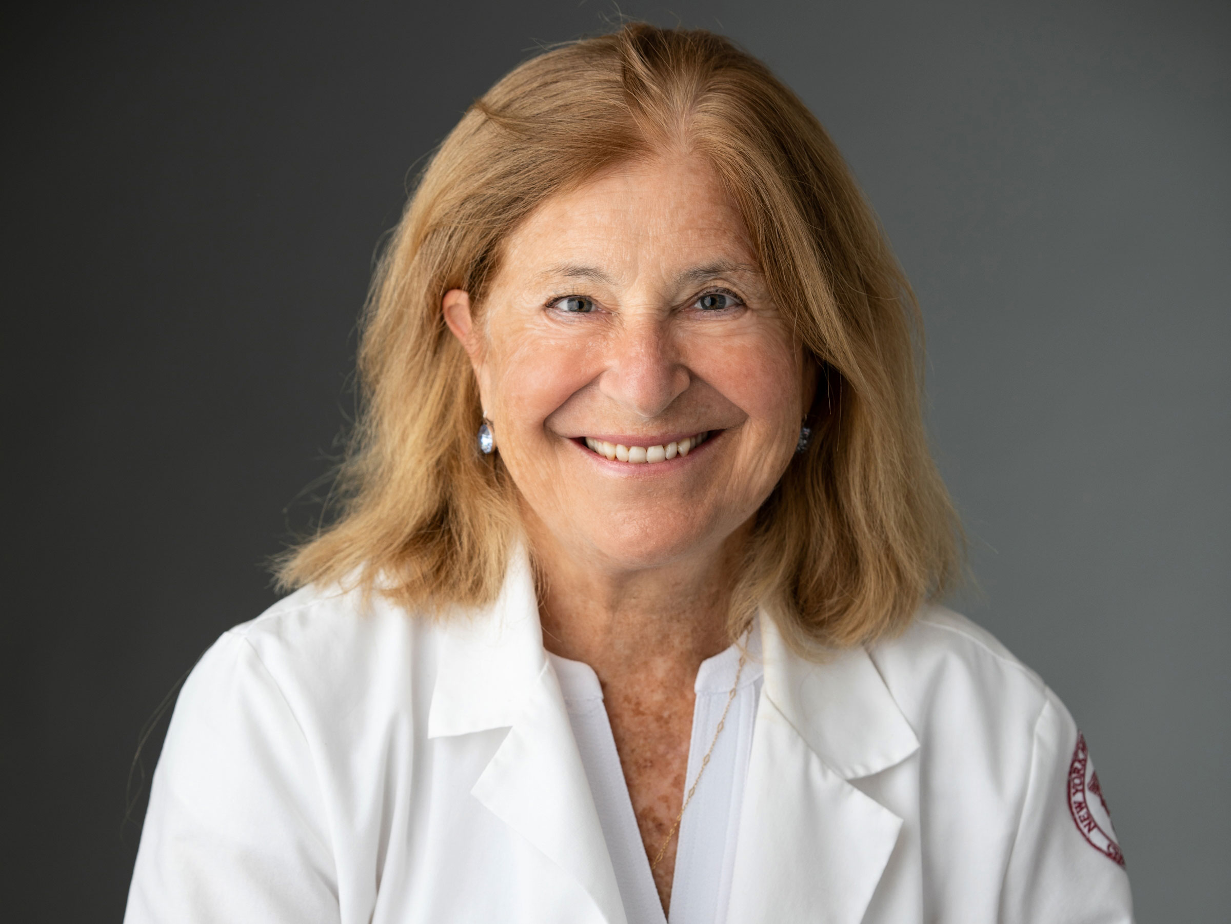 Female doctor smiling in front of a grey background with a white medical coat.