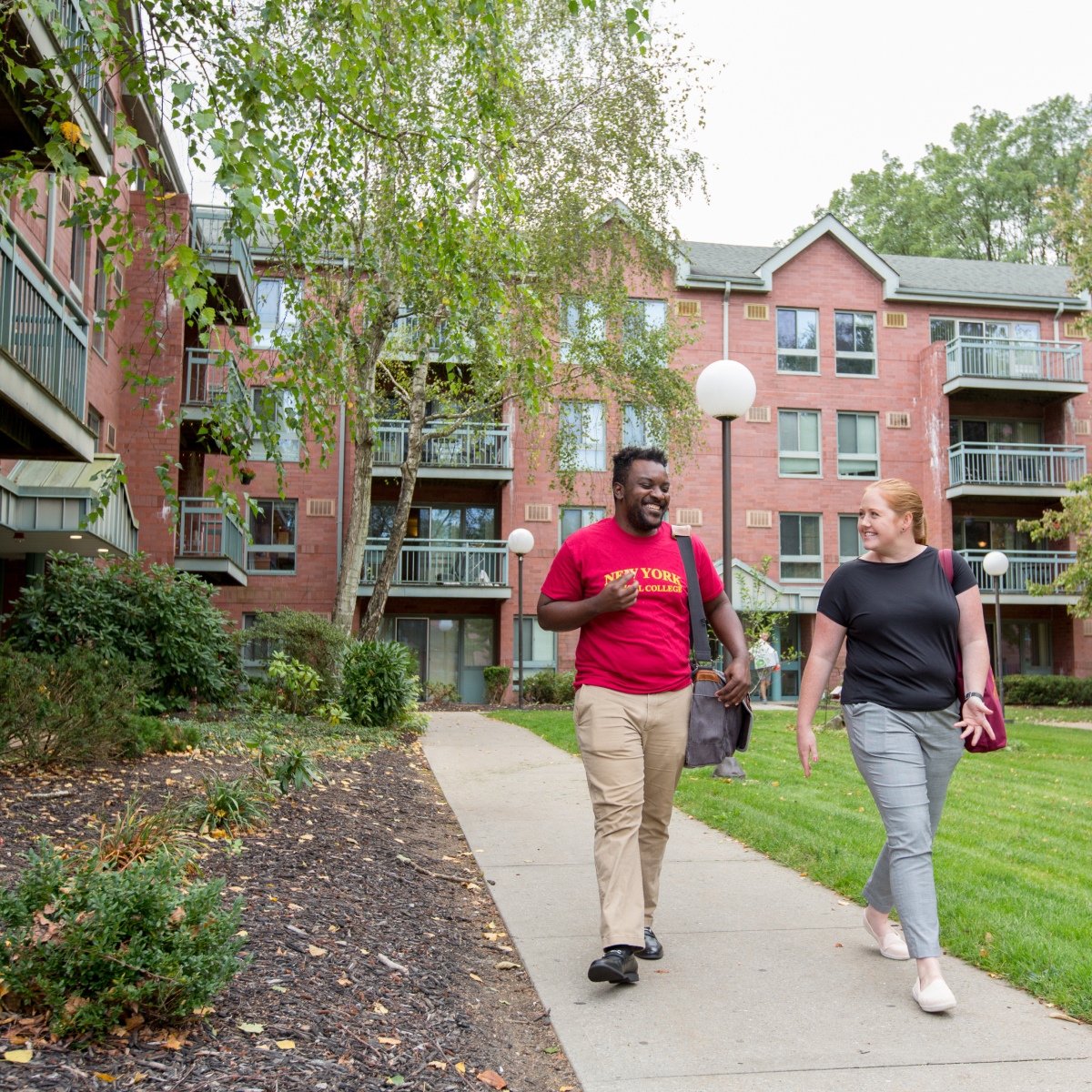 Two students walking on a path next to shrubbery outside a two-story dorm with balconies and a red brick facade