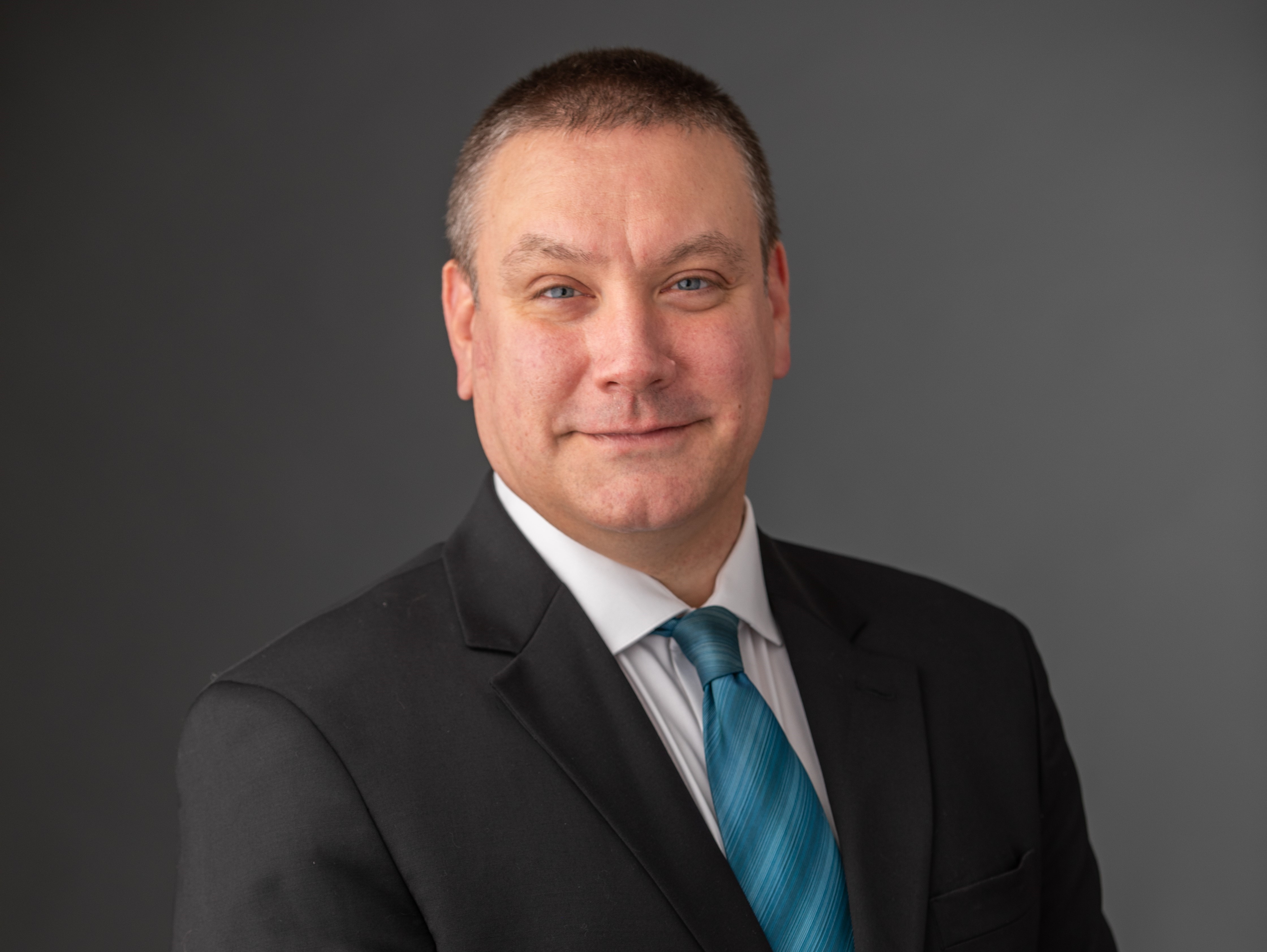 Man smiling in front of gray background smiling wearing a suit and tie