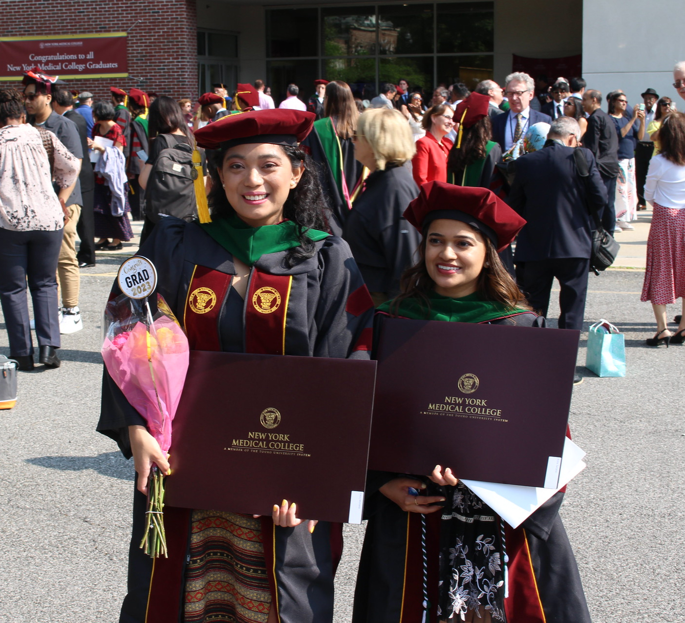 Outside their commencement ceremony, two graduates beam with smiles as they don their regalia, clutching diplomas and a bouquet of flowers